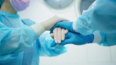 Ameliyat sonrası eldiven giyen doktorlar ameliyathanede meslektaşlarının yanında el ele tutuşuyorlar.