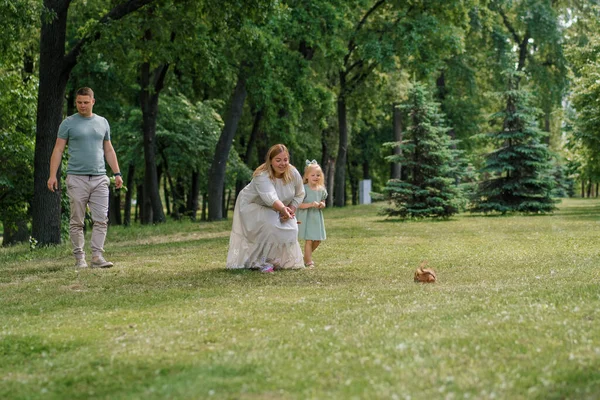 happy family on a walk in the park, mother shows her daughter a squirrel jumping on the grass child wants to feed it