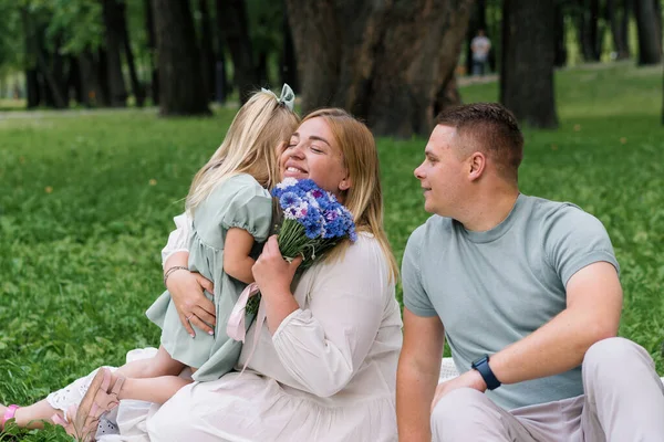 公園で幸せな家族は自然の中で休んでいます ドレスの小さな娘は彼女の母親に花束を与える 幸せな両親は娘にキス — ストック写真