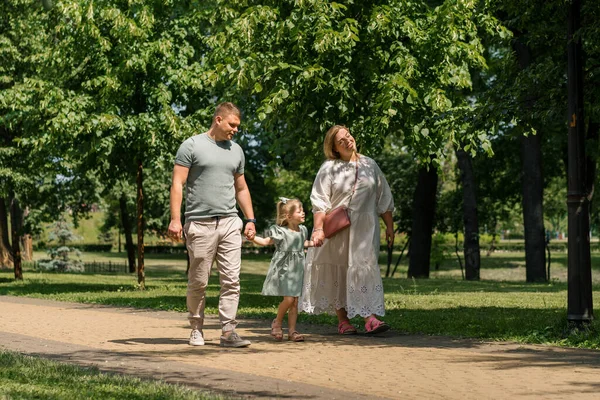 穿着绿色夏装的可爱小女孩 和父母一起在城市公园里散步夏季家庭生活幸福的家庭牵着手 — 图库照片