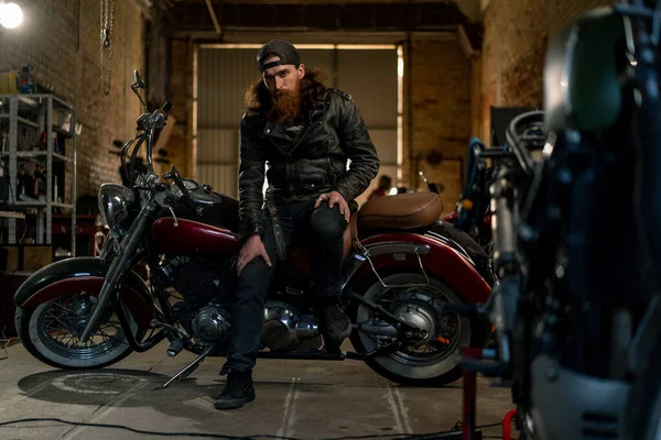 創造的な本物のオートバイの研修会のガレージの残酷なひげを付けられたバイカーのメカニックはオートバイで座っています — ストック写真