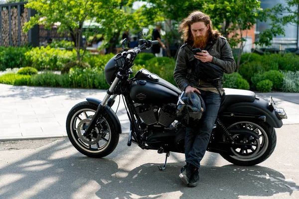 スピードレースの欲望の後 彼の手で携帯電話で黒いオートバイに座っている残酷な思慮深い赤毛のひげバイカー — ストック写真