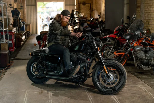 創造的な本物のオートバイの研修会のガレージの深刻な赤毛はバイカーのメカニックをオートバイで座らせました — ストック写真