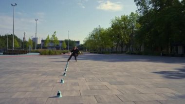 Parkın arka planında paten kayan sporcu kız sokak sporu konileri ile paten dersinin keyfini çıkarıyor.