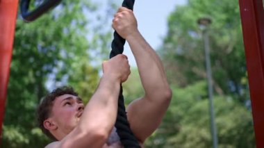 Çıplak gövdeli genç sporcu spor sahasındaki antrenman sırasında bir ipe tırmanıyor ve vücudunu şişiriyor.