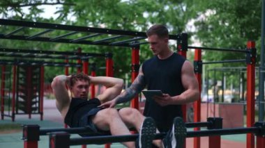 Elinde tabletle bir fitness eğitmeni genç bir sporcuyu spor sahasındaki motivasyon desteğiyle sokaklarda karın kaslarını şişiriyor.