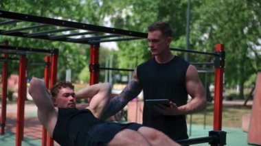 Elinde tabletle bir fitness eğitmeni genç bir sporcuyu spor sahasındaki motivasyon desteğiyle sokaklarda karın kaslarını şişiriyor.