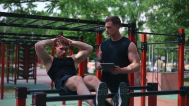 Elinde tabletle bir fitness eğitmeni genç bir sporcuyu spor sahasındaki motivasyon desteğiyle sokaklarda karın kaslarını şişiriyor.