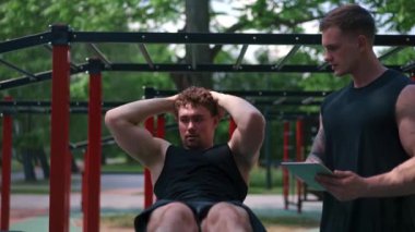 Elinde tabletle bir fitness eğitmeni genç bir sporcuyu spor sahasındaki motivasyon desteğiyle sokaklarda karın kaslarını şişiriyor.