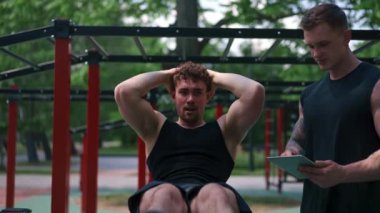 Elinde tabletle bir fitness eğitmeni genç bir sporcuyu spor sahasındaki motivasyon desteğiyle sokaklarda karın kaslarını şişiriyor.