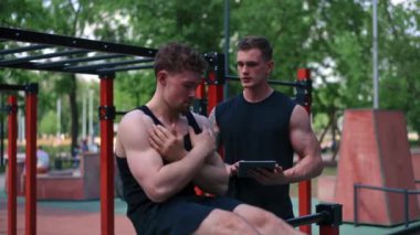 Elinde tabletle bir fitness eğitmeni genç bir sporcuyu spor sahasındaki motivasyon desteğiyle sokaklarda karın kaslarını şişiriyor.