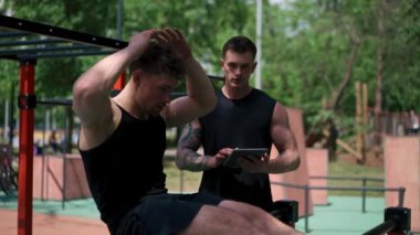 Elinde tabletle bir fitness eğitmeni genç bir sporcuyu spor sahasındaki motivasyon desteğiyle sokaklarda karın kaslarını şişiriyor.