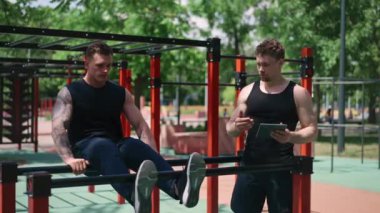 Elinde tabletle bir fitness eğitmeni genç bir sporcuyu spor sahasındaki motivasyon desteğiyle sokaklarda karın kaslarını şişiriyor.