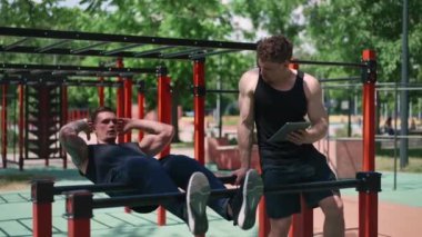 Elinde tabletle bir fitness eğitmeni genç bir sporcuyu spor sahasındaki motivasyon desteğiyle sokaklarda karın kaslarını şişiriyor.