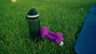 Su şişesi ve dambıllar parkta spor paspasının yanındaki çimlerin üzerine uzanıyorlar.