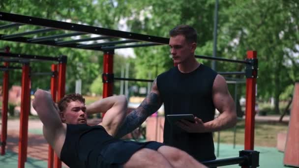 一个手里拿着石碑的健身教练训练一个年轻的运动员在街上用操场上的动力支撑着他的腹肌 — 图库视频影像