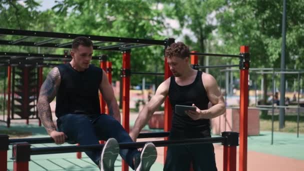 Trener Fitness Tabletem Ręku Trenuje Młodego Sportowca Pompującego Swoje Mięśnie — Wideo stockowe
