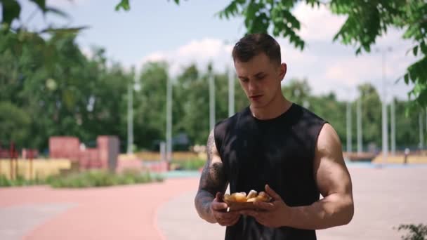 Молодий Спортсмен Після Тренування Тримає Солодкі Пончики Руках Вагається Їдять — стокове відео