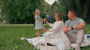 Mutlu bir aile parkta dinlenir, elbise giymiş küçük bir kız annesine bir buket çiçek verir mutlu bir anne baba kızını öper