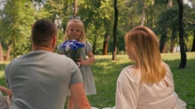Mutlu bir aile parkta dinleniyor. Elbise giymiş küçük bir kız annesine bir buket çiçek veriyor.