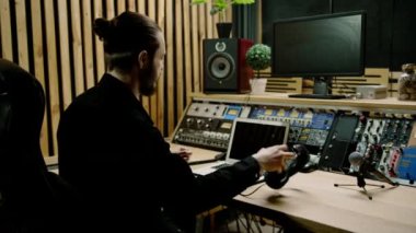 Genç ses mühendisi kulaklık takıyor. Profesyonel bir müzik stüdyosunda çalışıyor. Monitörleri var.