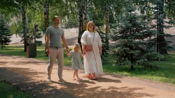 歩く夏の家族のライフスタイル 子供時代幸せな家族の手を握る公園の両親と緑の夏のドレスの小さなかわいい女の子 — ストック動画