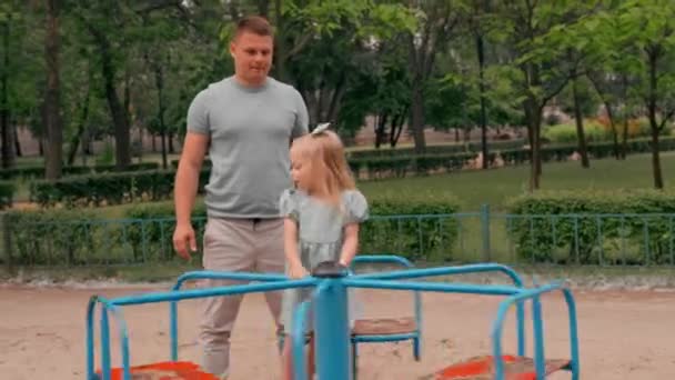 緑の夏のドレスの小さなかわいい女の子は 遊び場のお父さんに乗る娘のカルーセル夏の子供時代 — ストック動画