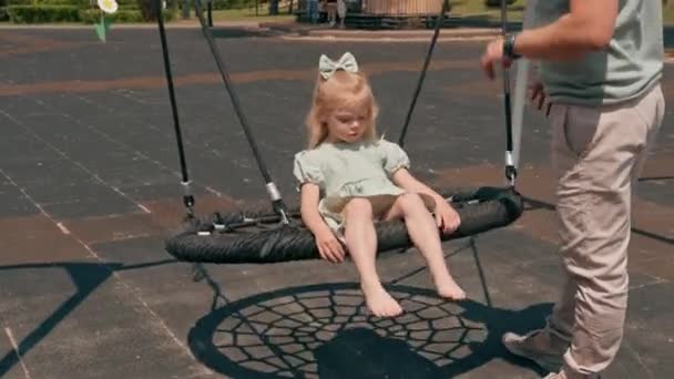 緑の夏のドレスの小さなかわいい女の子は 遊び場で彼女の頭に弓で 母親は夏の子供時代に彼女の娘に乗っています — ストック動画