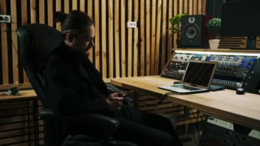 Genç bir ses mühendisi müzik stüdyosunda monitörlerle çalışıyor ve bir dengeleyici elinde bir akıllı telefon tutuyor telefonda bilgi arıyor