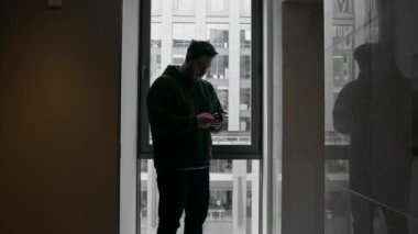Elinde telefon olan bir adam, iş merkezini gören bir pencerenin arka planında iş meselelerini çözer.