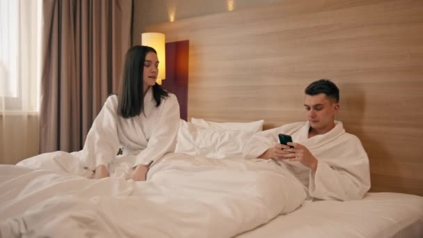 ホワイトローブのベッドに横たわるホテルの部屋のハネムーンの若いカップルの肖像画 紛争不満を論じる — ストック動画
