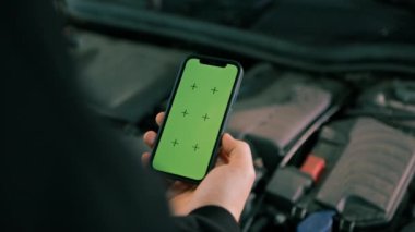 Elinde telefon olan ve lüks mavi arabanın kaputunun yanında yeşil ekran olan bir erkek araba servis elemanı.