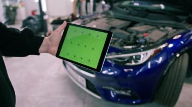 Elinde tabletle gezen erkek araba servisi çalışanı. Lüks mavi arabanın kaputunun yanında yeşil bir ekran var.