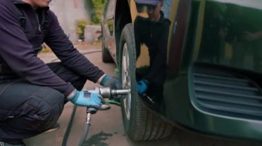 Araba tamircisi, siyah bir arabanın tekerini lastik dükkanındaki matkapla değiştirip araba tamiri yapıyor.