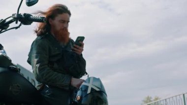 Vahşi, düşünceli, kızıl sakallı motosikletli, siyah motosikletli, elinde telefonla hız yarışından sonra elinde bir portre.