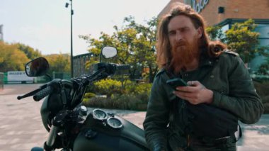 Vahşi, düşünceli, kızıl sakallı motosikletli, siyah motosikletli, elinde telefonla hız yarışından sonra elinde bir portre.