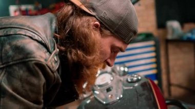 Yaratıcı otantik motosiklet atölyesi kızıl saçlı sakallı tamirci motosiklet garajında motosiklet koltuğunu tamir ederken yakın çekim aletleri kullanıyor.
