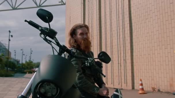 ひげを持つ残忍な赤毛のバイカーは レースの開始前に彼の手の渇きのスピードでヘルメットを保持する黒いオートバイに座っています — ストック動画