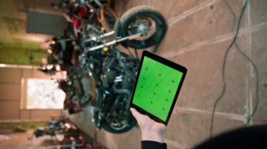 Garajda motorsiklet tamircisi. Motosikletin yanında duruyor ve elinde yeşil ekran tableti var.