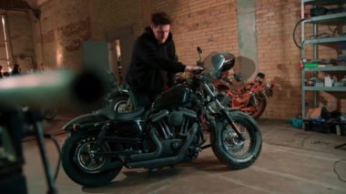 Motosiklet atölyesinde siyah kapüşonlu ciddi sakallı bir motosikletli teftiş yapıyor.