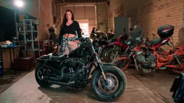 Otantik motosiklet atölyesi güzel genç motorcu kız havalı siyah motosikletin yanında duruyor.