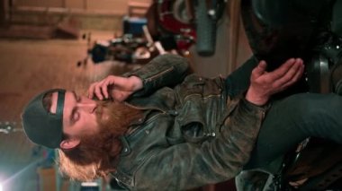 Otantik motosiklet atölyesi garajı portresi ciddi kızıl saçlı sakallı motorcu motosiklet tamircisi motosiklette oturup telefonda dikey video konuşuyor.