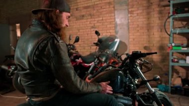 Yaratıcı otantik motosiklet atölyesi garajı portresi ciddi kızıl sakallı motorcu motosiklet tamircisi oturuyor.