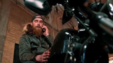 Otantik motosiklet atölyesi garajı portresi ciddi kızıl sakallı motosikletli tamirci motosiklette oturuyor ve telefonla konuşuyor.