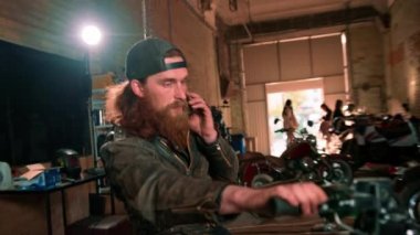 Otantik motosiklet atölyesi garajı portresi ciddi kızıl sakallı motosikletli tamirci motosiklette oturuyor ve telefonla konuşuyor.