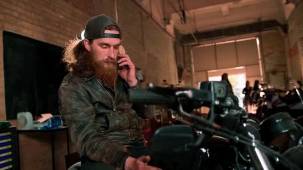 真实的摩托车修车厂车库肖像严肃的红头发 留着胡子的摩托车技师坐在摩托车上 讲电话 — 图库视频影像