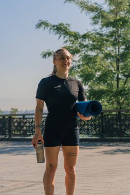Parkta antremana başlamadan önce elinde bir su şişesi ve kettlebell tutan gülümseyen genç bir sporcu kızın portresi.