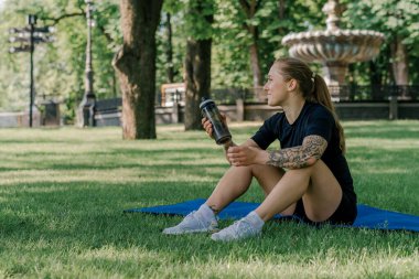 Spor minderinde dinlenen genç bir kızın portresi. Parkın sağlıklı yaşam tarzı konseptini öğrendikten sonra açık havada su içmek.