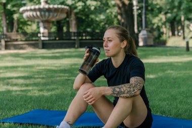 Spor minderinde dinlenen genç bir kızın portresi. Parkın sağlıklı yaşam tarzı konseptini öğrendikten sonra açık havada su içmek.