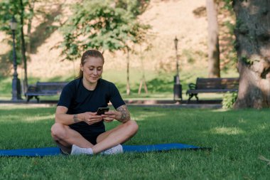 Spor minderinde, elinde akıllı bir telefonla parkta dinlenen genç bir bayan sporcunun portresi. Sağlıklı yaşam tarzı kavramı.